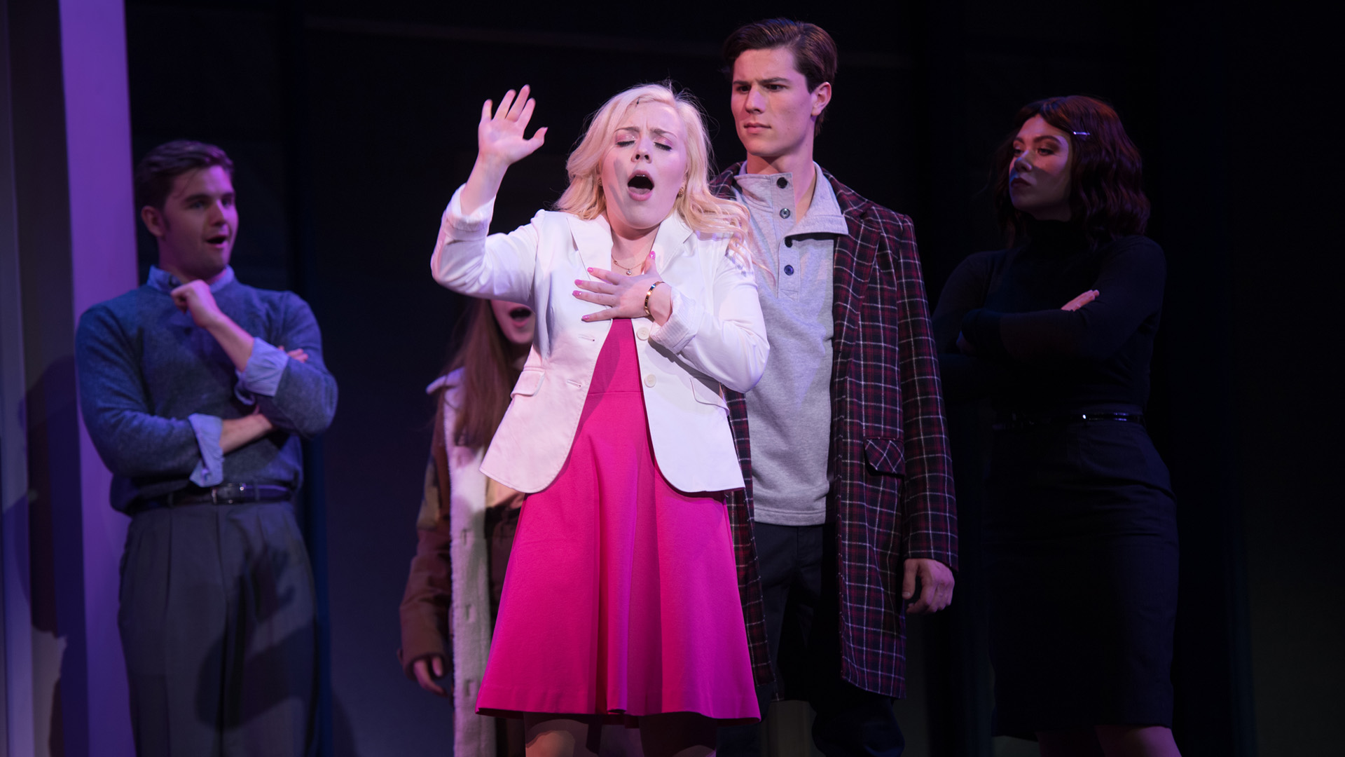 Student performers act and sing on stage during a musical theatre production, wearing costumes and using expressive movement under theatrical lighting.